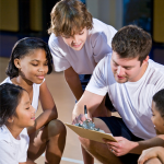 young students surrounding a coach who is showing them instructions on a clipboard